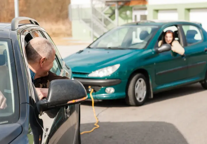 Mężczyzna w czarnym aucie patrzy na kobietę w zielonym samochodzie, połączonym żółtym kablem.