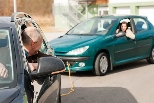 Mężczyzna w czarnym aucie patrzy na kobietę w zielonym samochodzie, połączonym żółtym kablem.